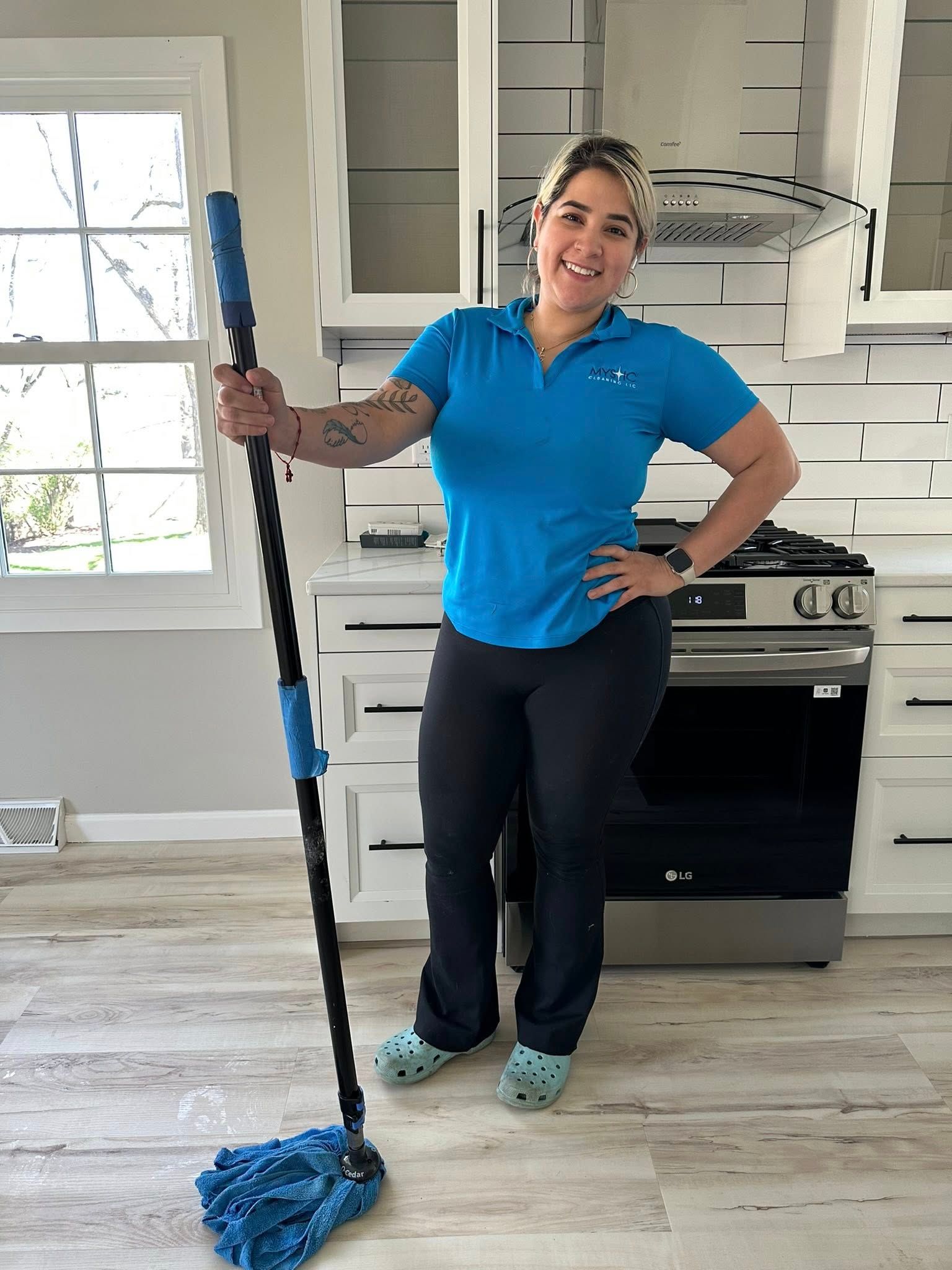 Woman holding a mop in a kitchen, smiling. Wearing a blue shirt, black pants, and teal clogs.