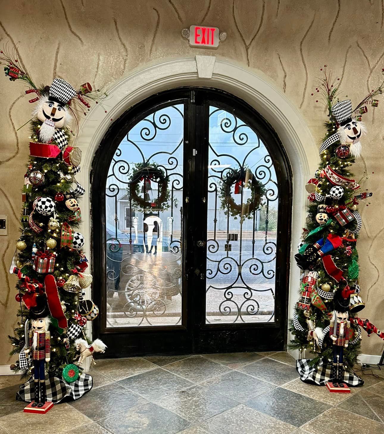 Two christmas trees are in front of a door with an exit sign above it.