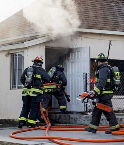 Firefighters entering a building engulfed in smoke, one carrying a chainsaw and dragging a hose.