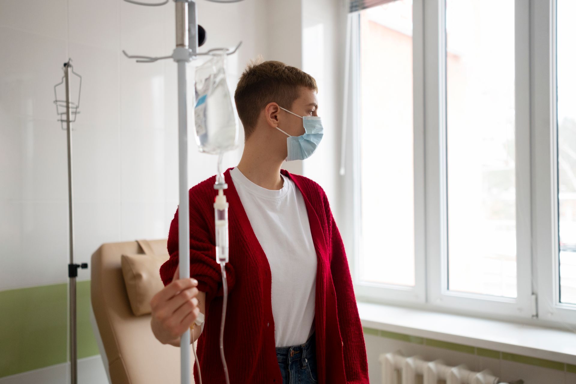 A woman wearing a mask is standing next to a drip in a hospital room.