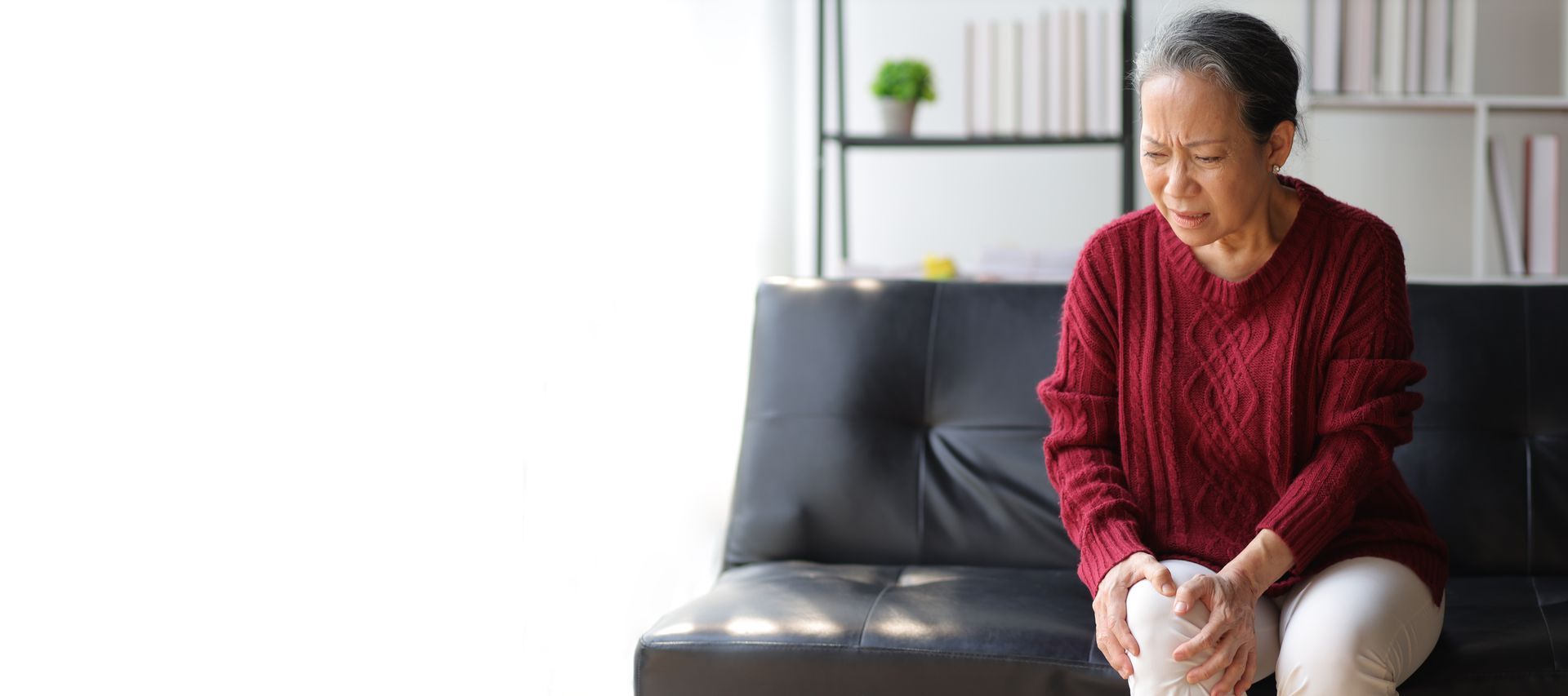 An elderly woman is sitting on a couch holding her knee in pain.