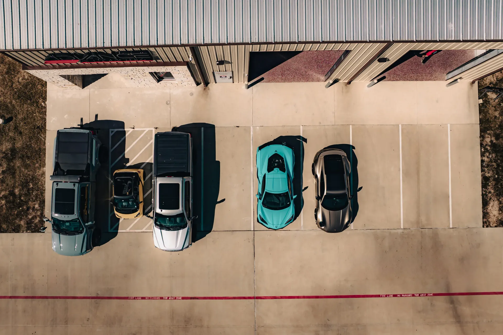 Overhead view of several cars parked outside a building: two trucks, a small black car, and two sports cars, one blue, one gray. | Advance Automotive