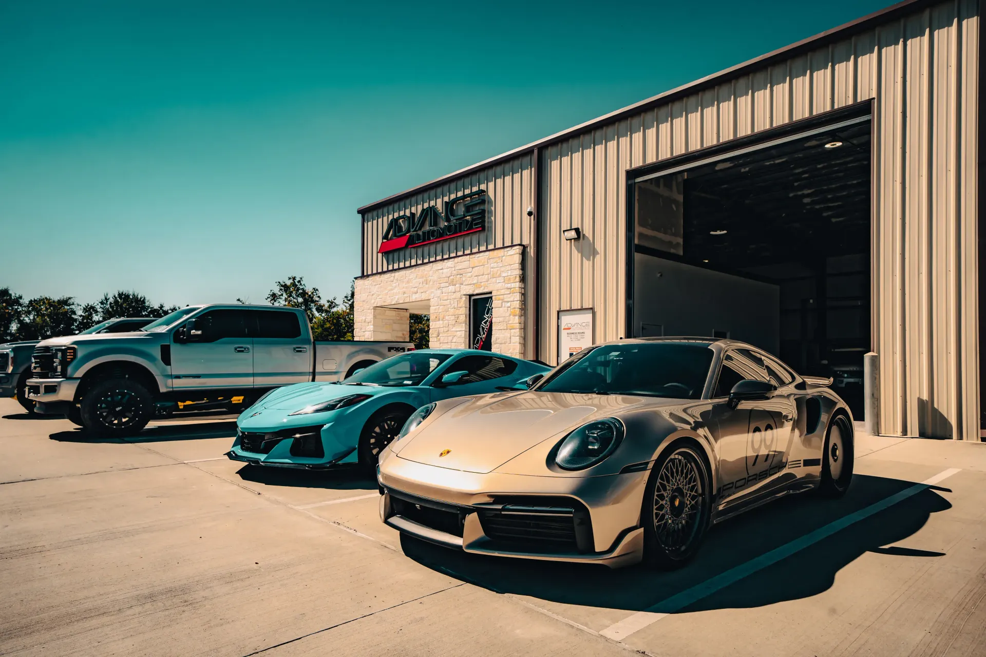 Silver Porsche and blue Corvette parked outside a garage, with a silver truck in the background on a sunny day. | Advance Automotive