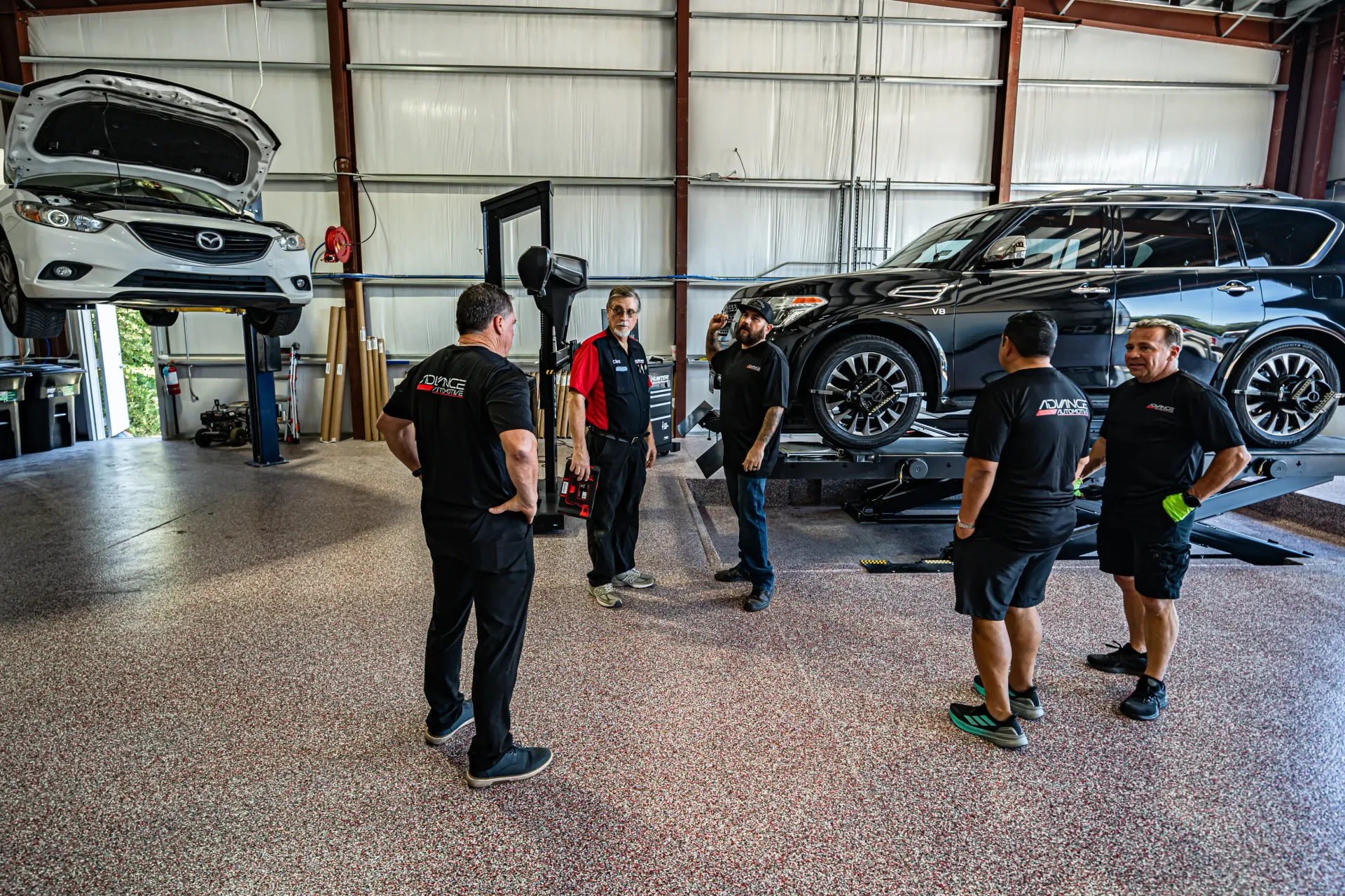 Five mechanics in a car garage with two cars on lifts, discussing a repair. | Advance Automotive