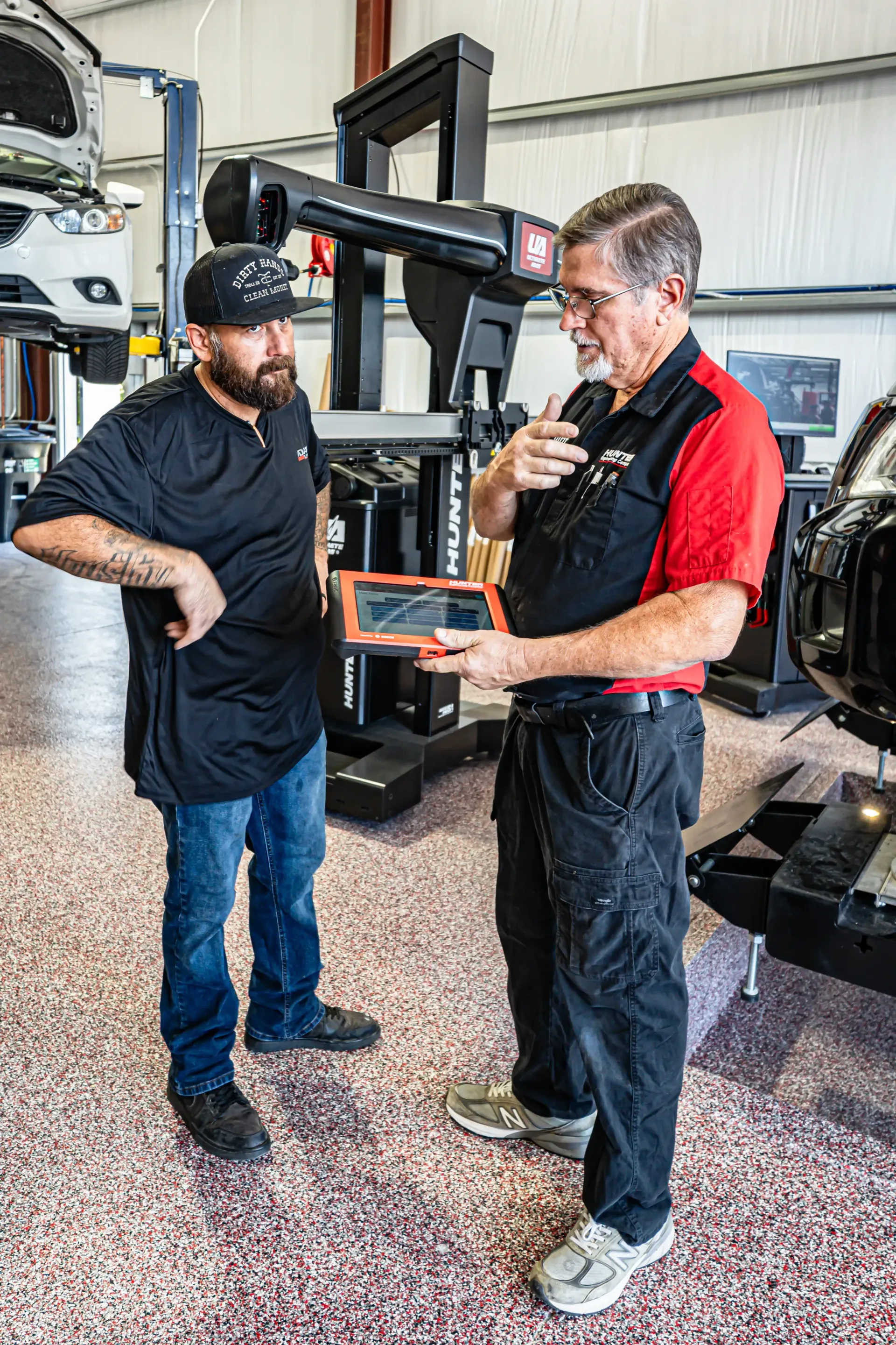 Two men in auto shop; one holding a tablet. Man in cap and jeans listens as other in red shirt gestures. Alignment machine in background. | Advance Automotive
