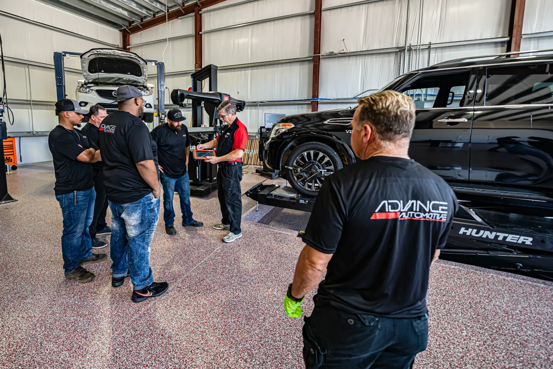 Group of people in a garage around a car on a lift; someone points at the car with the Advance Auto logo. | Advance Automotive
