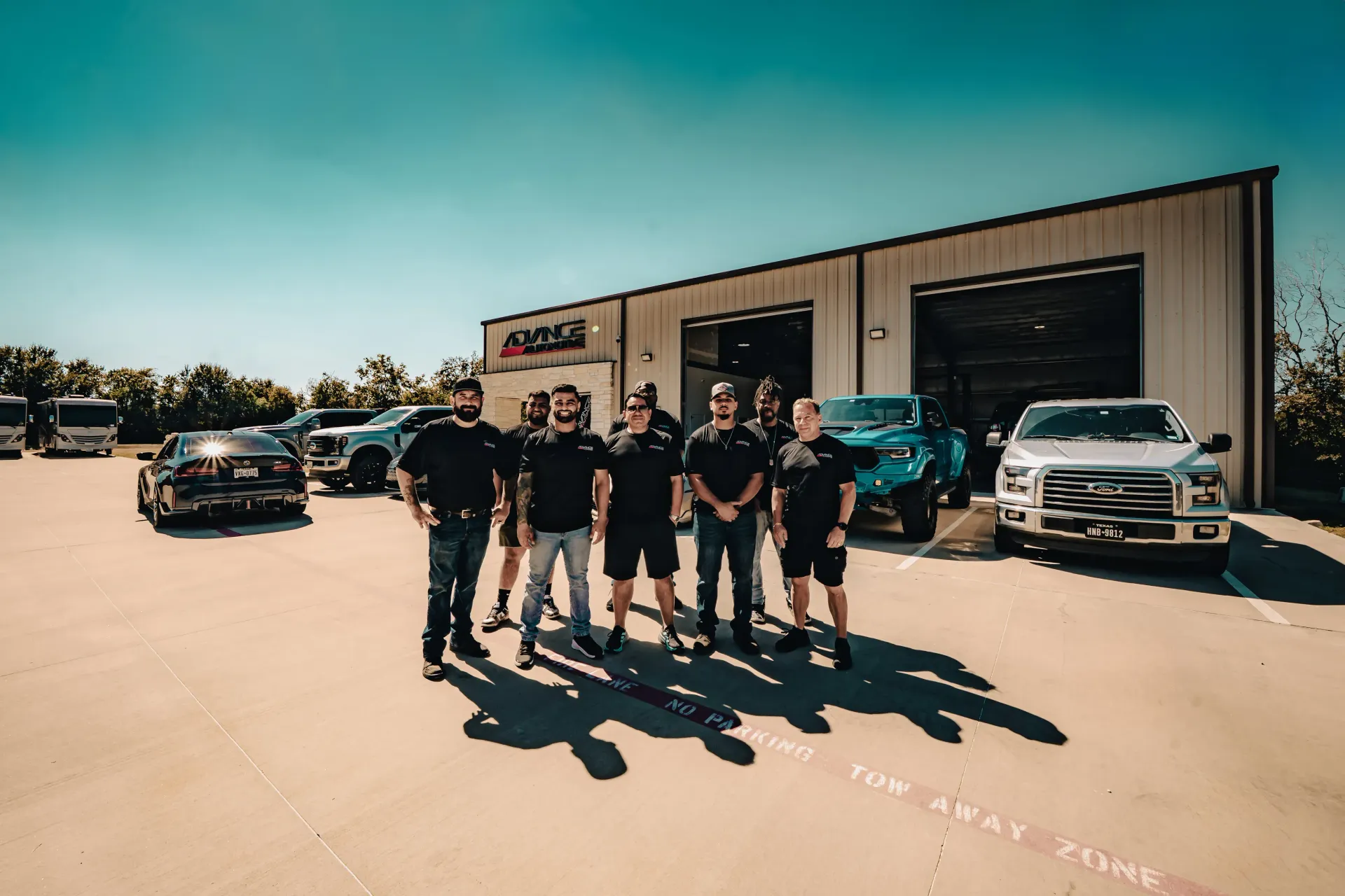 A group of people stand in front of a building with cars parked in a sunny parking lot. | Advance Automotive