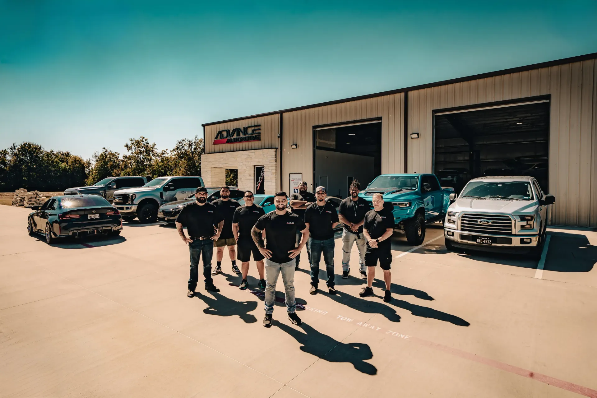 Group of people posing in front of a shop with several vehicles parked outside. | Advance Automotive