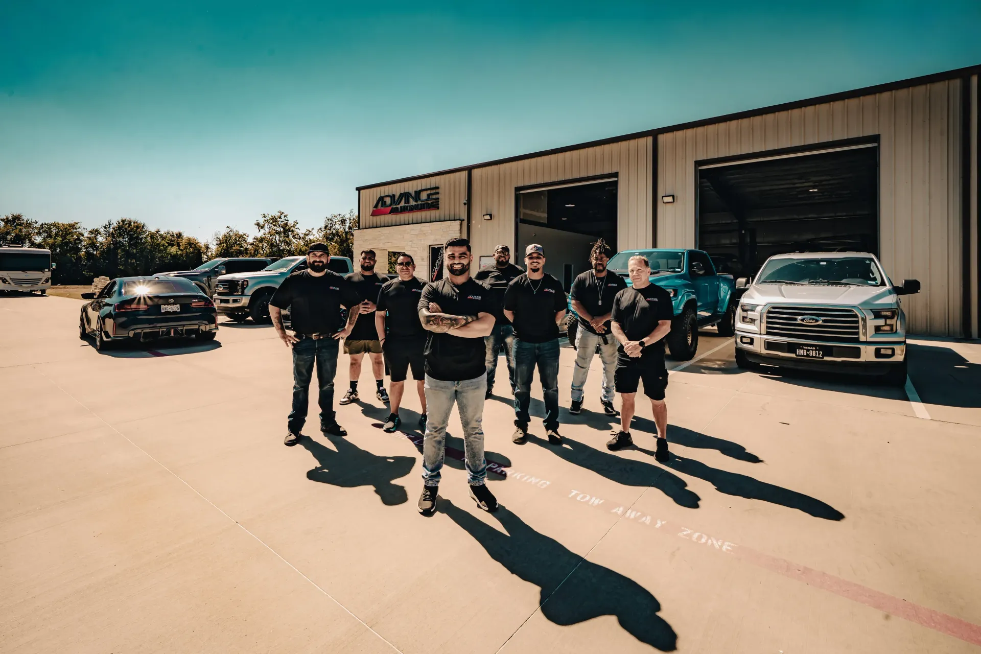 Team of men in front of a garage with cars. Sunny day, all wearing black shirts, arms crossed. | Advance Automotive