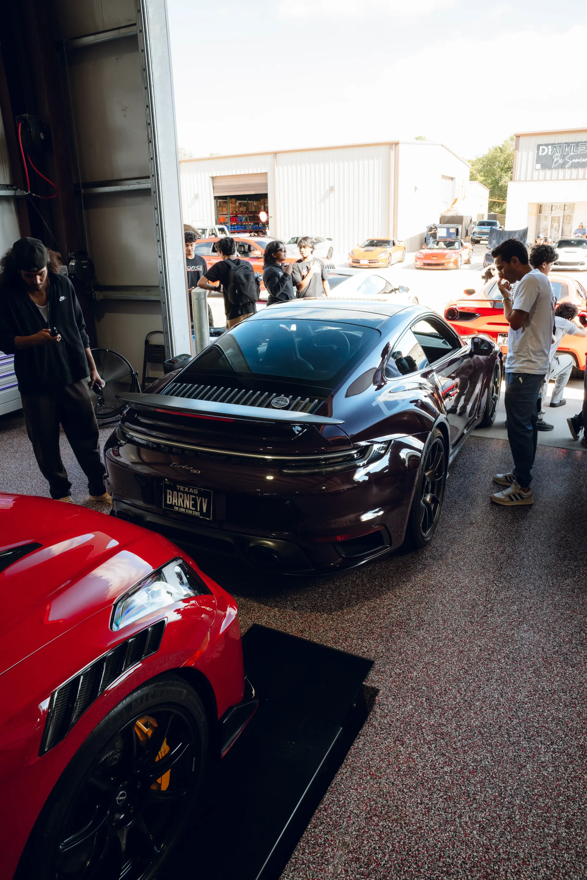 Dark purple Porsche sports car on display, red car in foreground, people milling around in a garage. | Advance Automotive