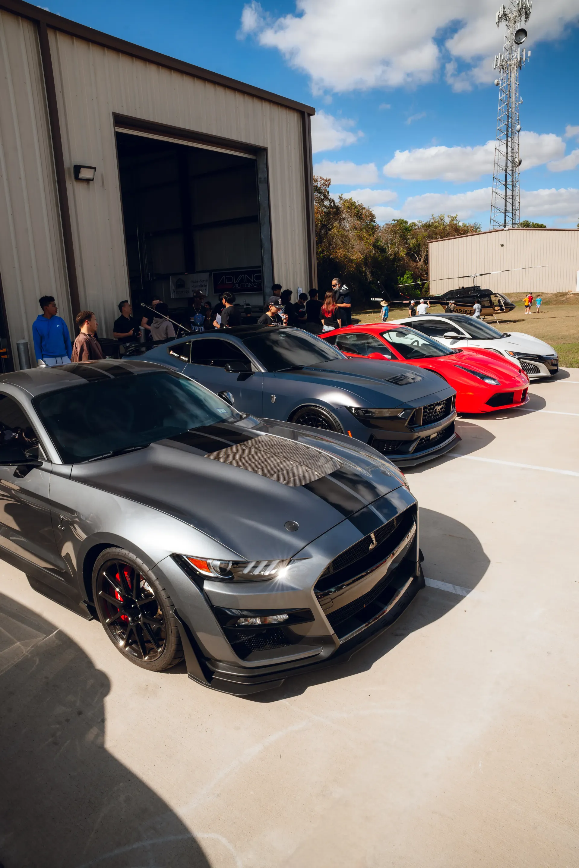 Lineup of sports cars parked outside a building on a sunny day. A crowd of people is visible in the background. | Advance Automotive
