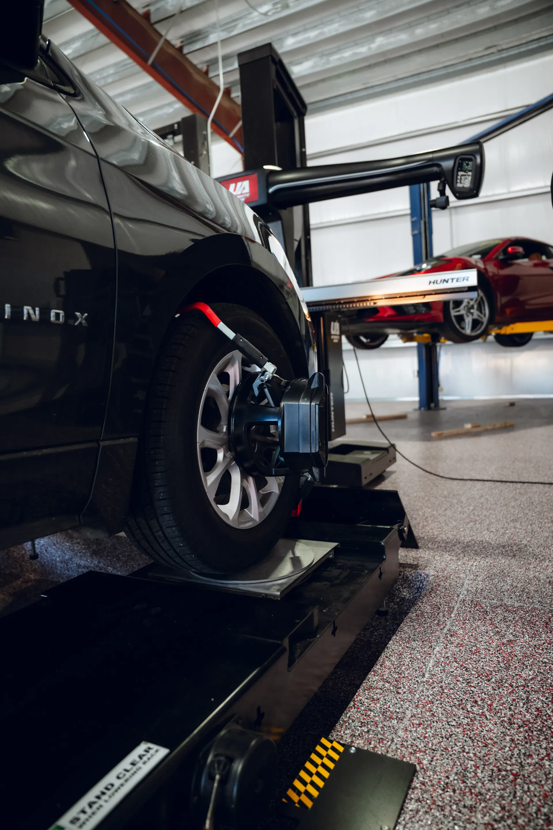 Black SUV undergoing wheel alignment in an auto shop. Red car on a lift in the background. | Advance Automotive