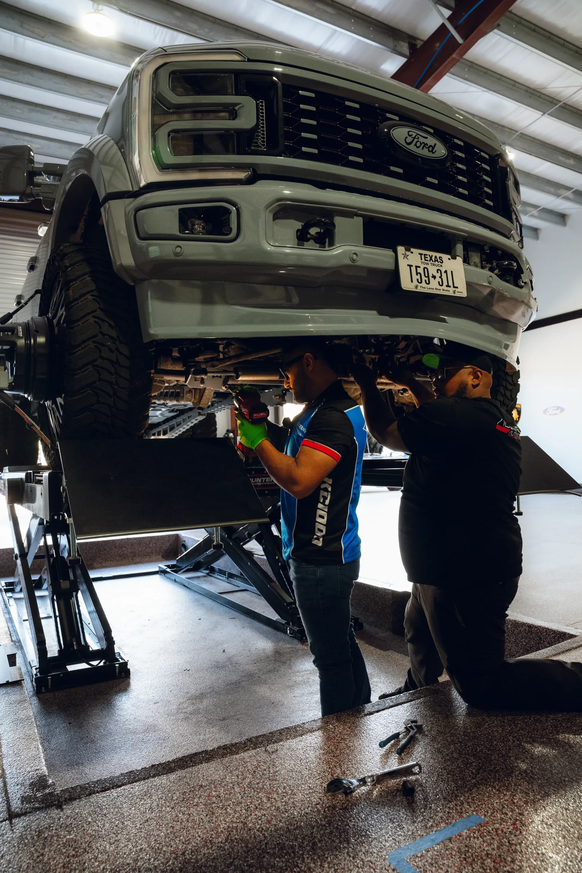 Two mechanics working on a gray Ford truck raised on a lift in a shop. | Advance Automotive