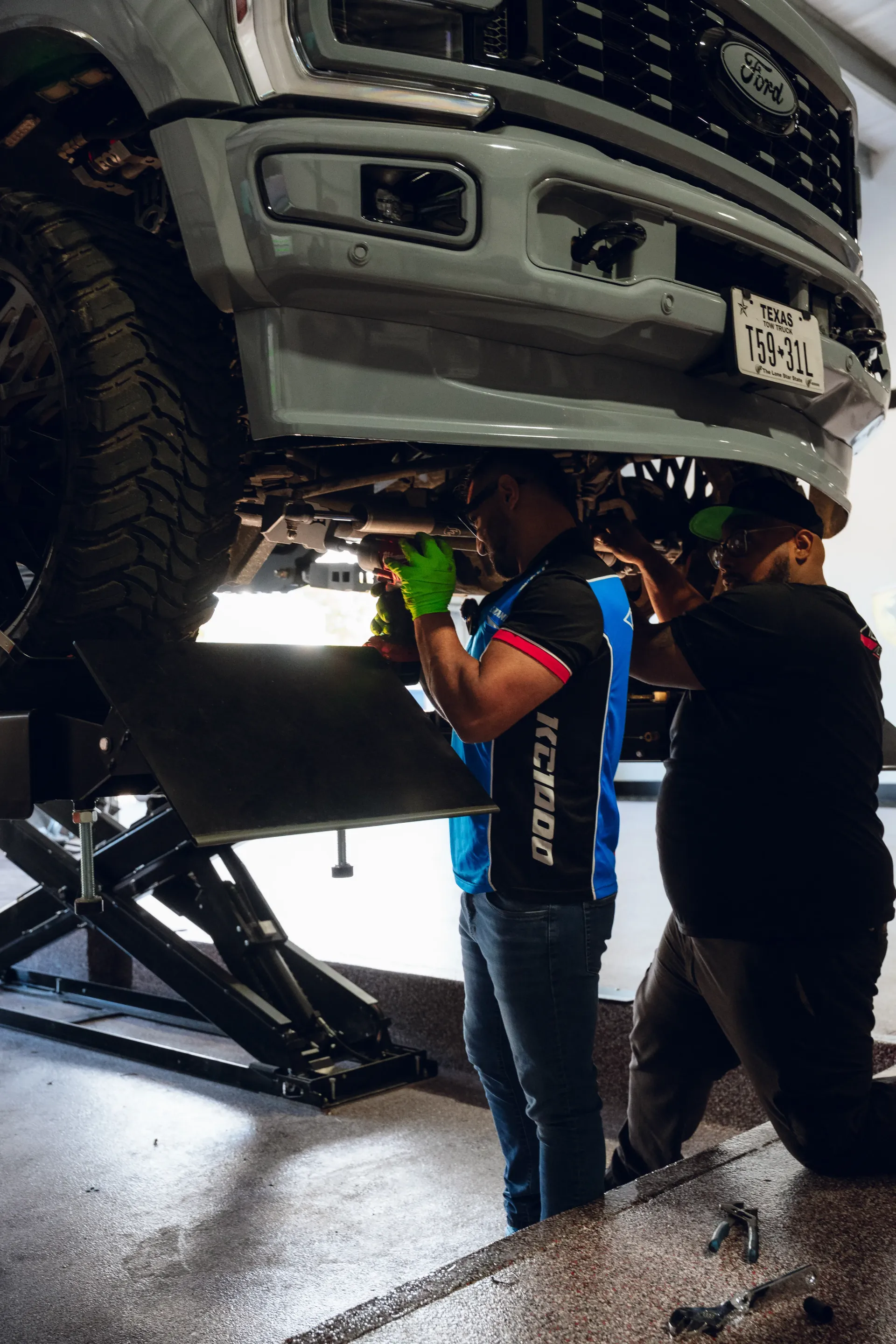 Two mechanics work under a gray truck on a lift, wearing gloves and blue shirts. | Advance Automotive