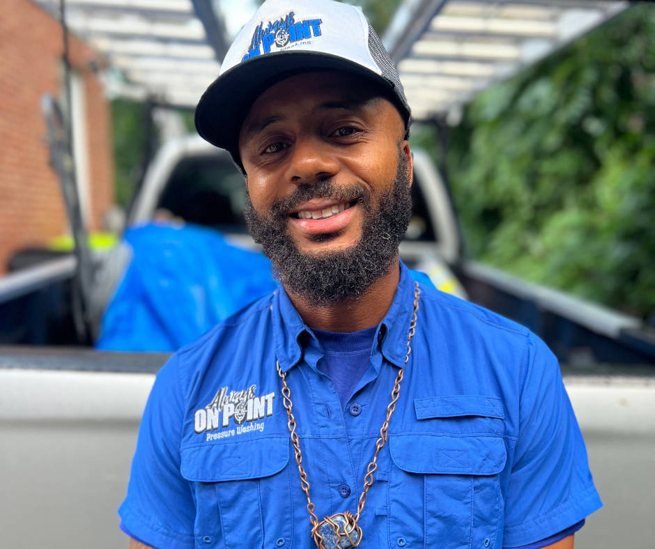 Smiling Black man in blue shirt and trucker hat, in front of a truck, outdoors.