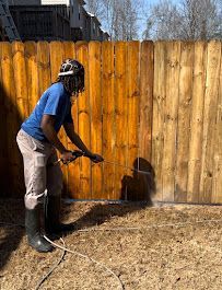 A person in a blue shirt and grey pants pressure washes a wooden fence outdoors, creating a misty spray.