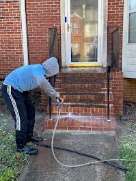 Person wearing a hood and sweats power washes brick steps leading to a home's front door.