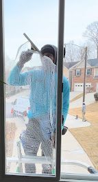 Person cleaning a window with a squeegee. They wear a blue shirt, gray pants, and a black cap.  