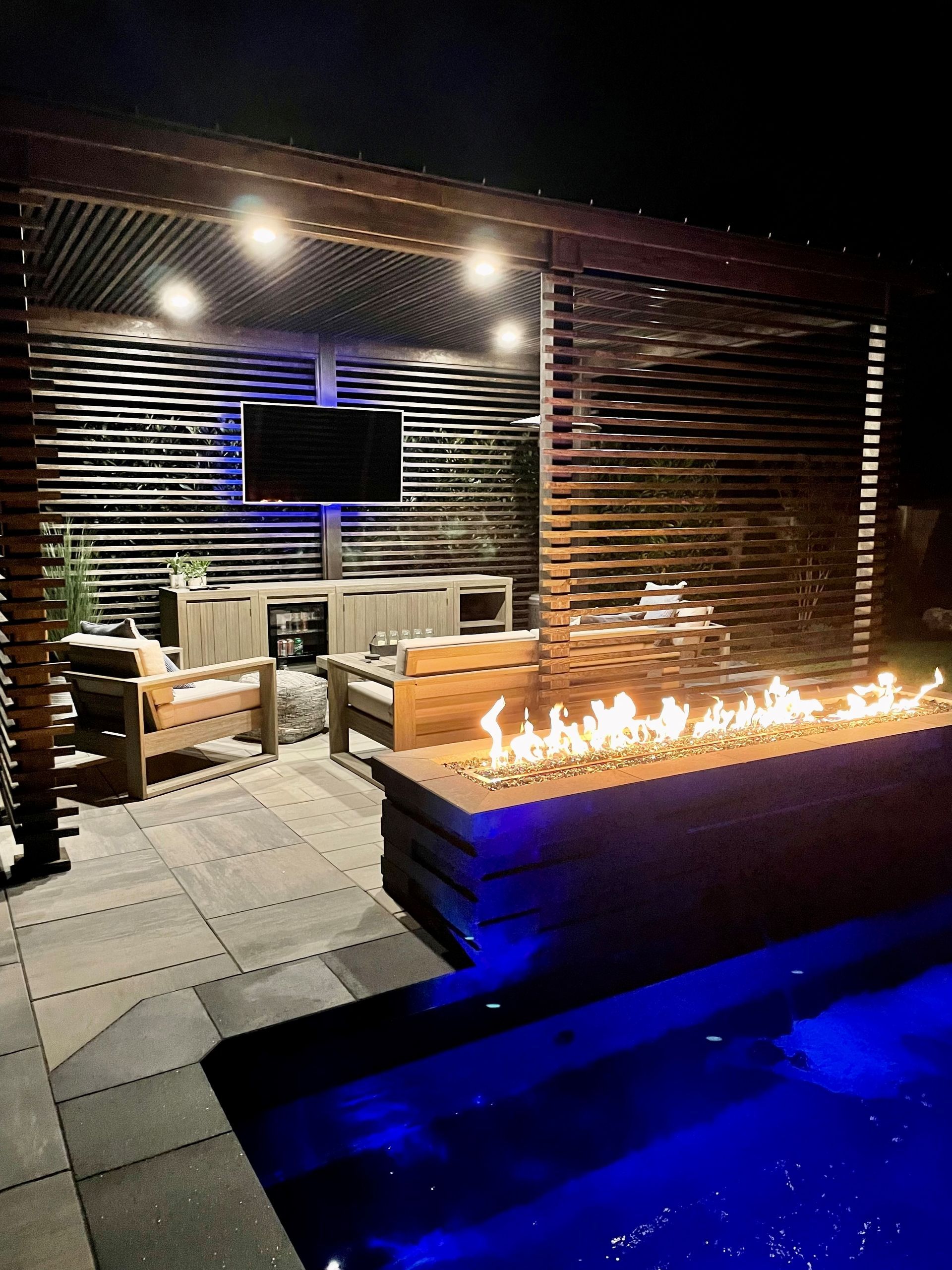 Night scene of a backyard patio with pool, fire pit, seating area, TV, and pergola with string lights.