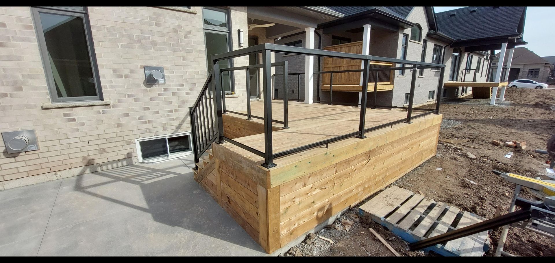 Wooden deck with black railings attached to the side of a light brick house, on a construction site.