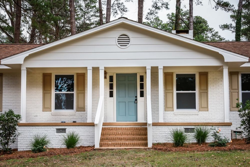 Front view of a beige house with a light blue door, white trim, and brick steps among pine trees