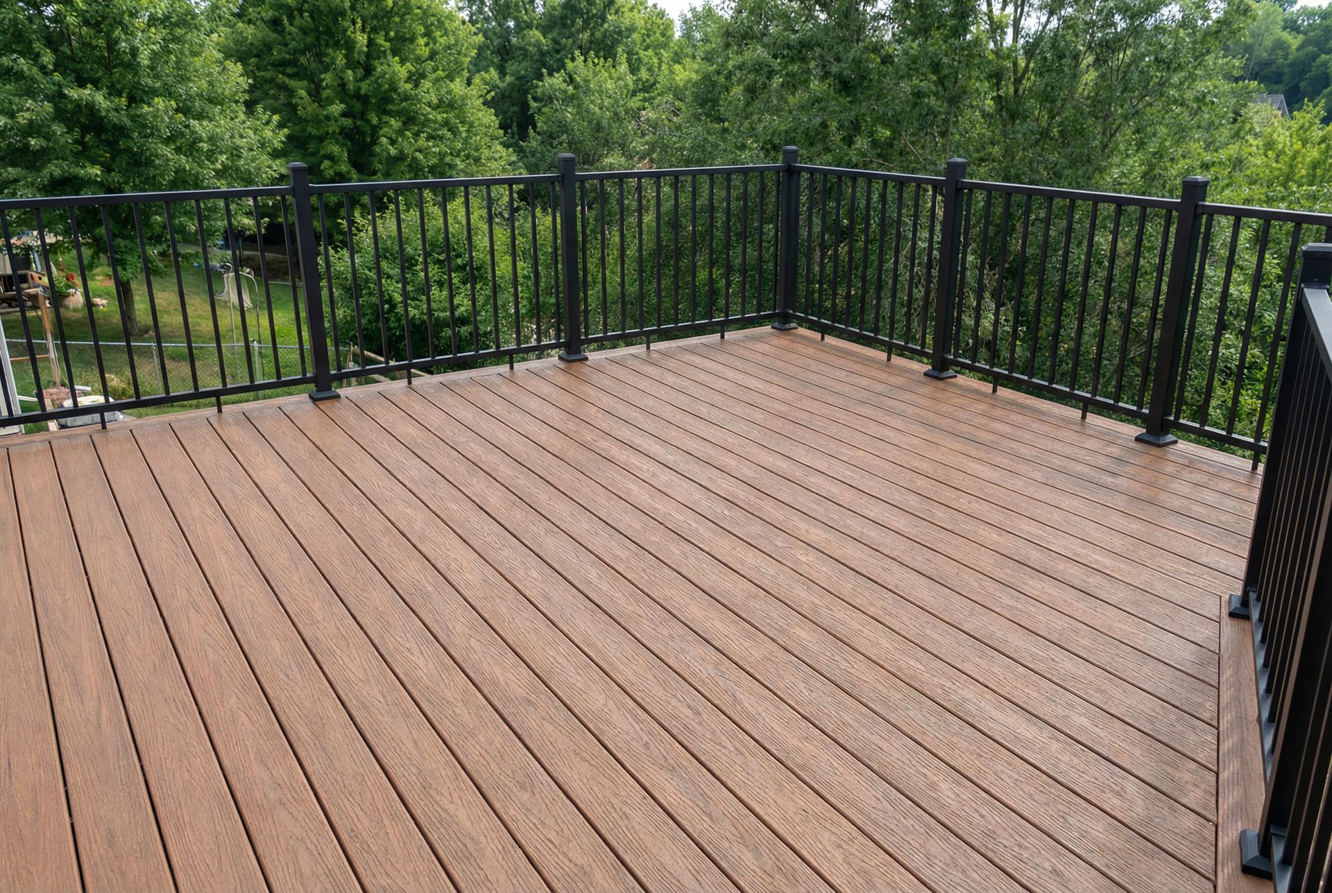 A brown composite deck with black metal railing, surrounded by green trees.