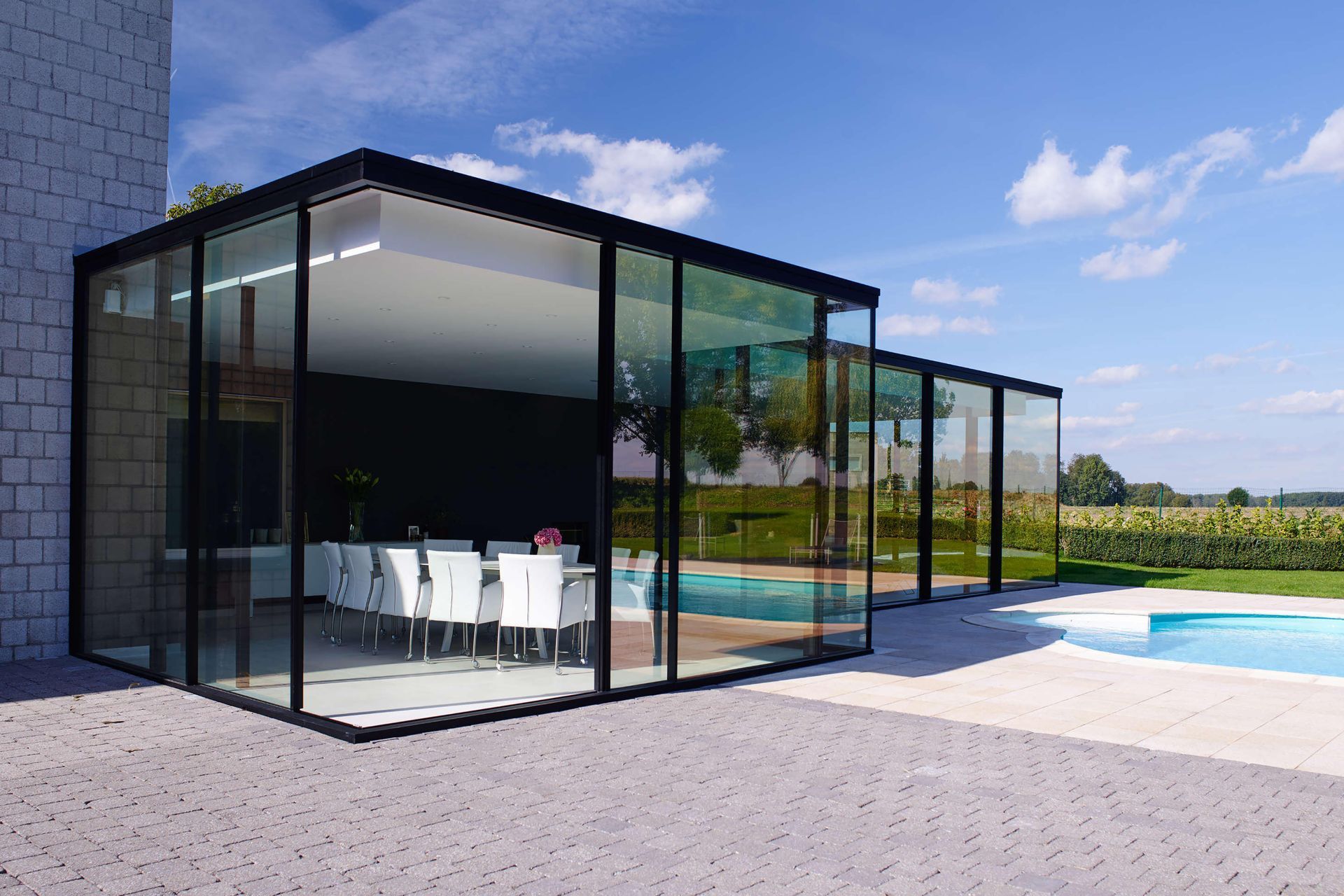 A large glass building with a dining table and chairs next to a swimming pool.