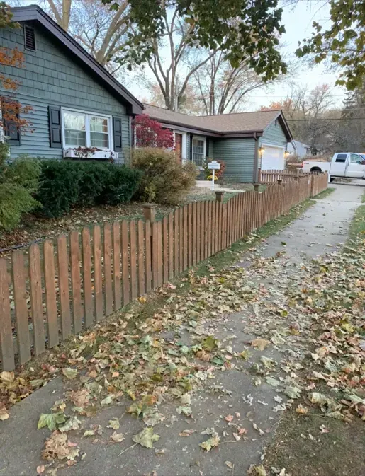 A wooden picket fence surrounds a house with leaves on the sidewalk.