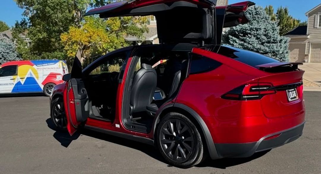 A red tesla model x with its doors open is parked in a driveway.