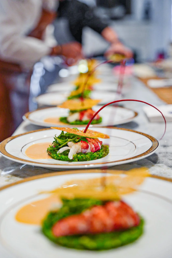 Plated gourmet food being prepared in a kitchen. Green base topped with lobster, crisp wafer, and red garnish.