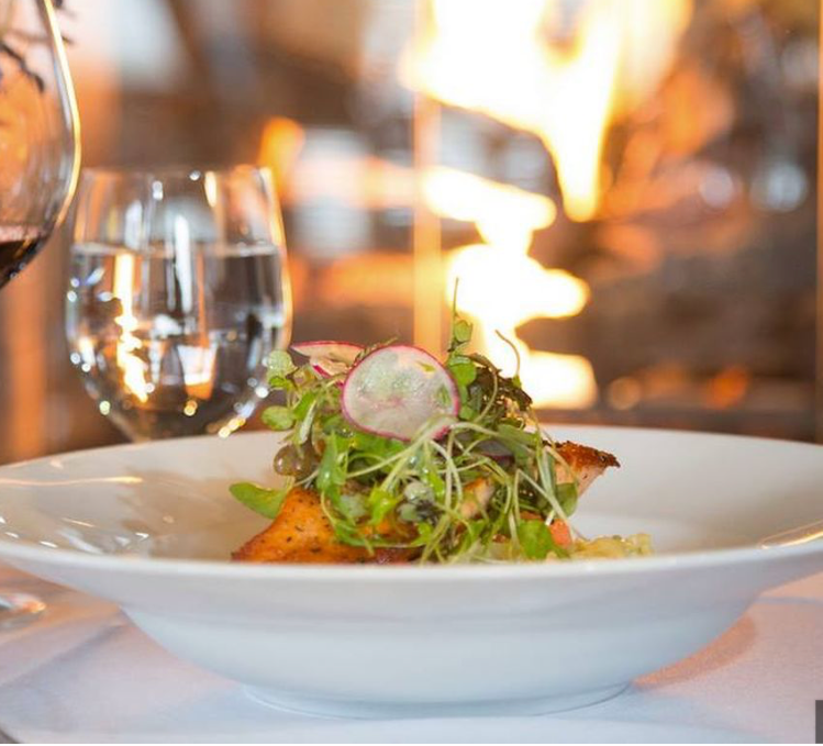 Salmon dish with greens, radish slices, and a glass of wine near a fireplace.