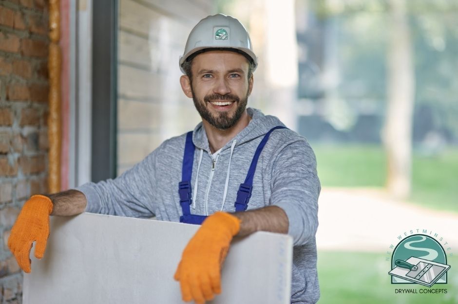 A drywall repair company worker smiles brightly, wearing a white hard hat, blue overalls, and a gray hoodie. He is holding a white drywall sheet and wearing bright orange work gloves. Commercial construction site background with brick walls visible near E 27th Ave, and Rupert St, South Vancouver, BC.