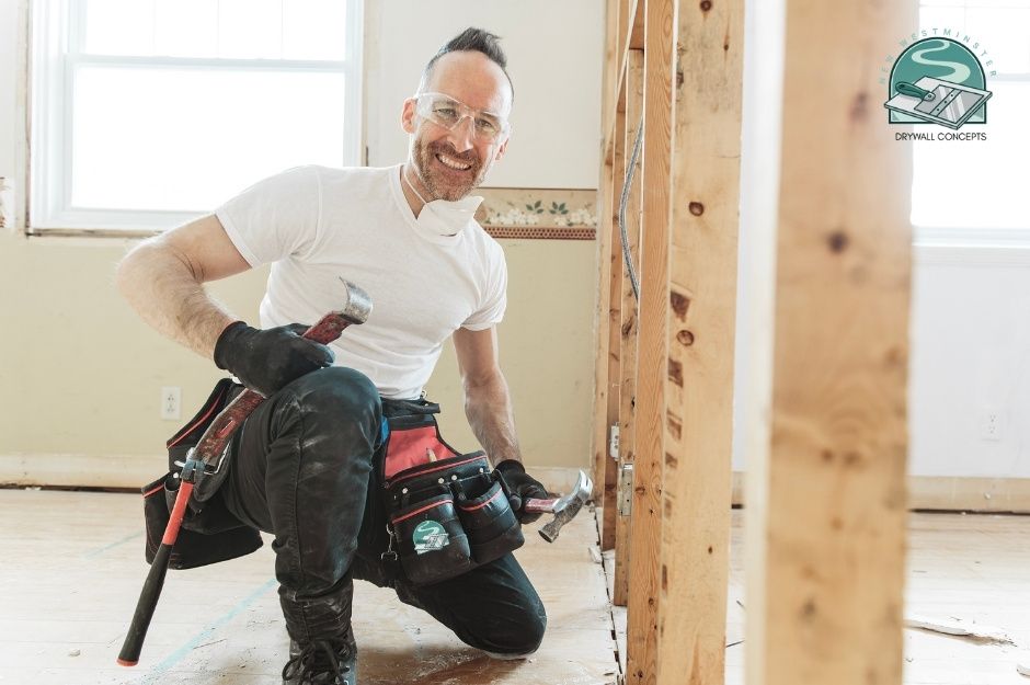 A drywall repair contractor kneels smiling on a commercial project site, holding a hammer. He wears a white t-shirt, safety glasses, and a tool belt. Exposed wooden framing and unfinished drywall are visible in the construction interior near Baycrest Ave, and Monson Cres, Coquitlam, BC.