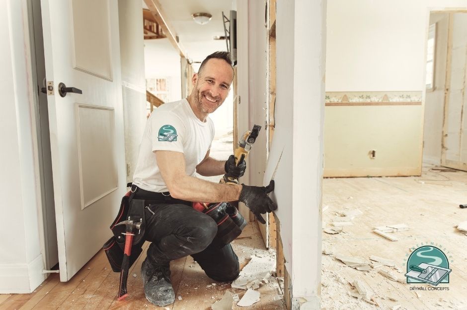 A smiling drywall repair contractor kneels in a home renovation area, holding a reciprocating saw while working on wall framing. Construction debris are scattered on the wooden floor around white walls and doorways in this residential project near 52 Ave, and 203 St in Langley, BC.