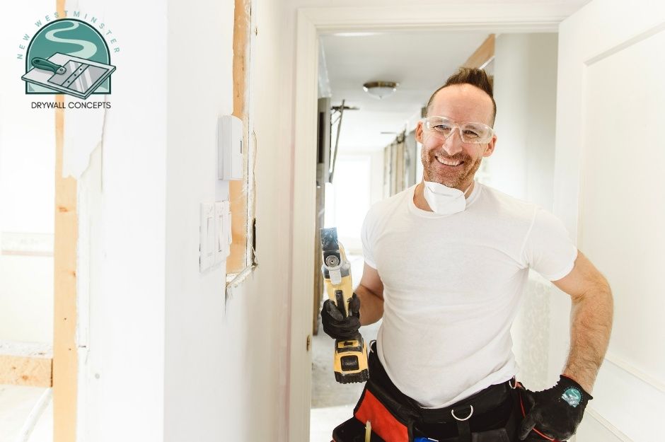 A drywall repair contractor smiles while holding a yellow oscillating multi-tool in a residential construction site hallway. He wears safety glasses, a white t-shirt, and a tool belt. Exposed wooden framing and white drywall are visible along the walls near Riverwood Gate, and Amazon Dr, Port Coquitlam, BC.