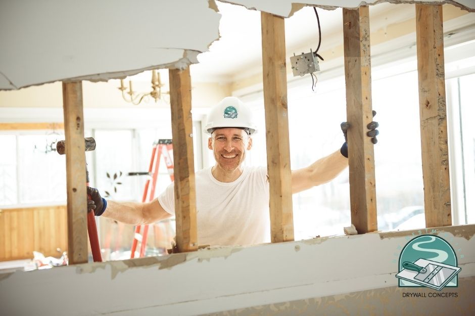 A drywall repair contractor wearing a white hard hat and t-shirt smiles while working on exposed wooden framing inside a residential construction site. He stands among vertical wood studs with damaged drywall and ceiling visible near Blackwood St, and Thrift Ave, White Rock, BC.