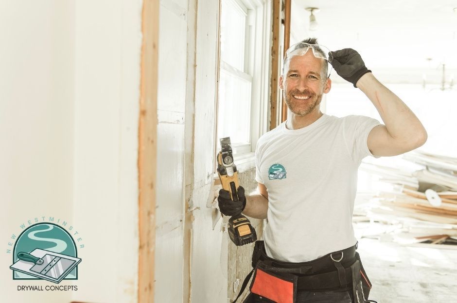 A drywall repair contractor smiles while working at a commercial construction site, wearing a white t-shirt and a tool belt, holding a cordless drill in black gloves. He stands among white drywall and wooden framing near 208 St, and 84 Ave, Willoughby, BC.