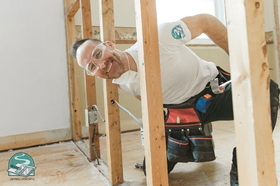 A drywall repair handyman in safety glasses and white shirt smiles while kneeling and working with wooden framing studs in an unfinished residential interior. He wears a tool belt and repairs framework near Amble Greene Dr, and 20 Ave, Surrey, BC.