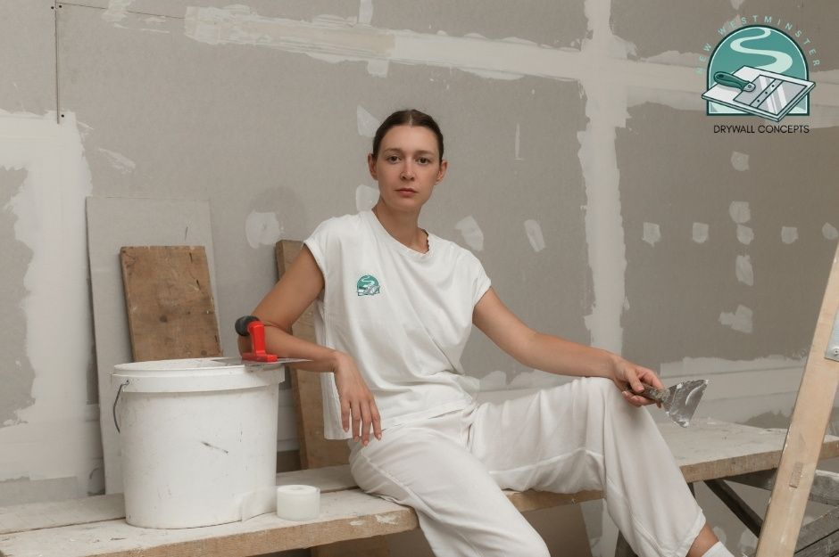 A drywall repair handyman in white clothing sitting on a wooden bench in an unfinished room, holding a putty knife tool, with a paint bucket nearby and patched drywall walls visible behind her, demonstrating repair work in a commercial construction site near Marguerite St, and W 37th Ave, Vancouver, BC.