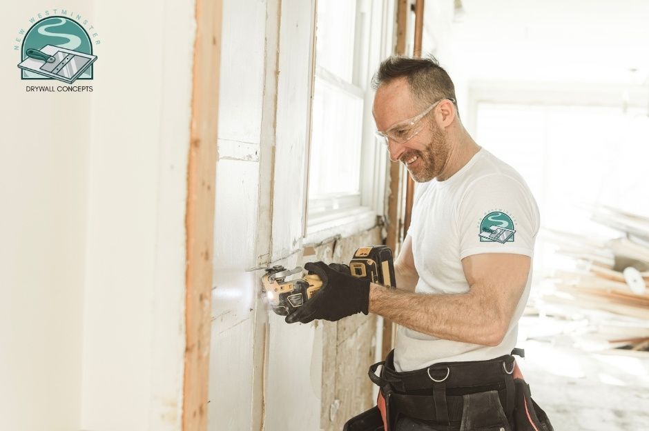 A drywall repair specialist, wearing safety glasses and black gloves, operates an oscillating multi-tool at a commercial construction site. He is smiling while working, wearing a white t-shirt and a tool belt in a room with exposed framing near 136 Ave, and 232 St, Maple Ridge, BC.