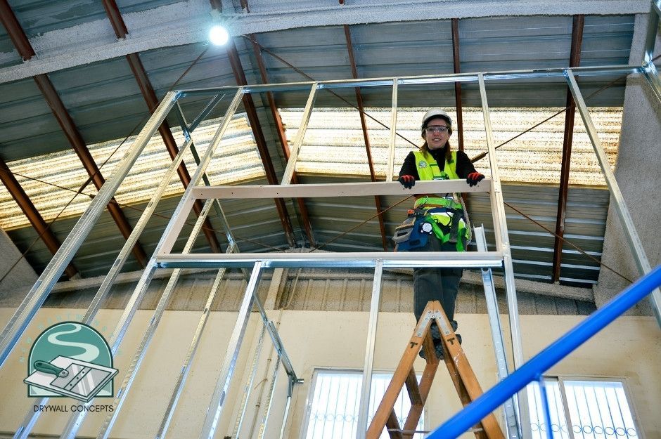 A drywall repair specialist in high-visibility safety gear and hard hat stands on a wooden stepladder, installing metal stud framing for drywall in a commercial building with exposed ceiling beams and concrete walls near Rutland Rd, and Caulfeild Dr, West Vancouver, BC.
