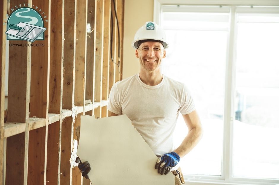 A drywall repair technician wearing a white hard hat and t-shirt, smiling while holding a large piece of drywall with both hands. He is standing in a residential construction site with exposed wooden wall studs and bright windows behind him, located near 44 Ave, and 50 St, Delta, BC.