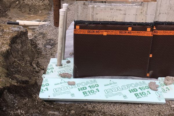 Mur de fondation avec isolation et membrane de drainage; tuyaux enterrés visibles.