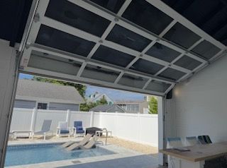 Open glass garage door revealing a backyard pool. White fence, lounge chairs, and sunlight.