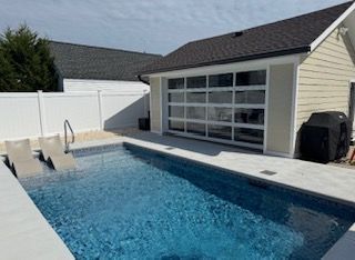 Pool with clear glass garage-style door leading into a light beige building. Poolside has two lounge chairs.