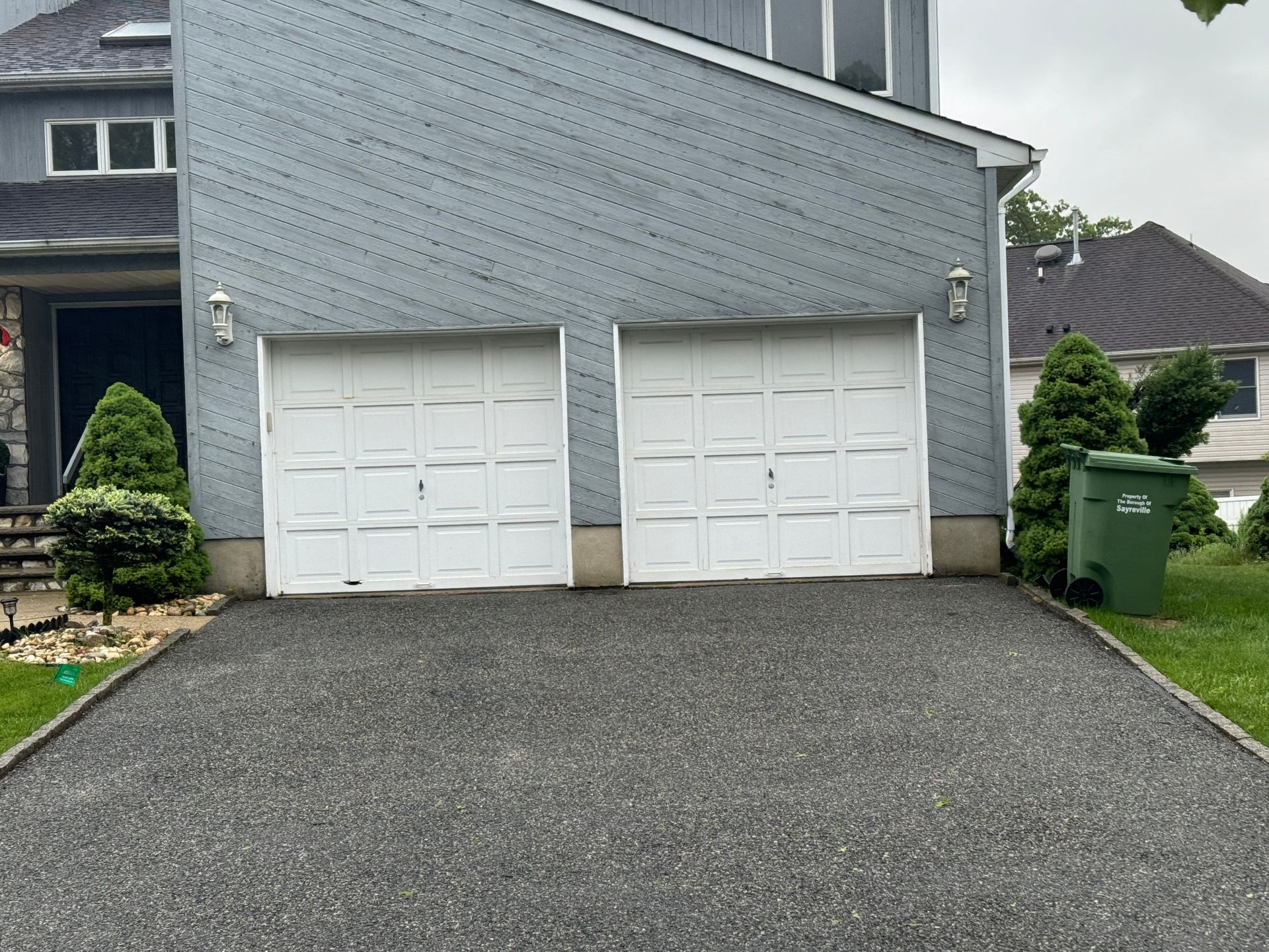 A house with two garage doors and a green trash can in front of it.