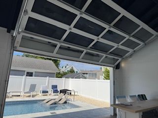 Open glass garage door reveals pool area with chairs and fence.