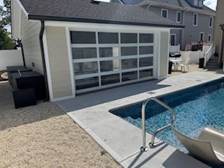 Garage with glass door next to a pool. Beige siding, black trim, and blue water are visible.