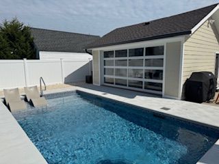 Pool with lounge chairs, next to a building with a glass garage door, and a grill.
