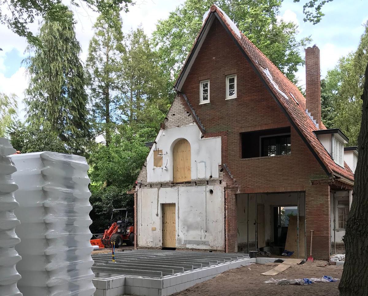 House under renovation, brick and wood exterior, red roof, surrounded by trees. Construction materials visible.